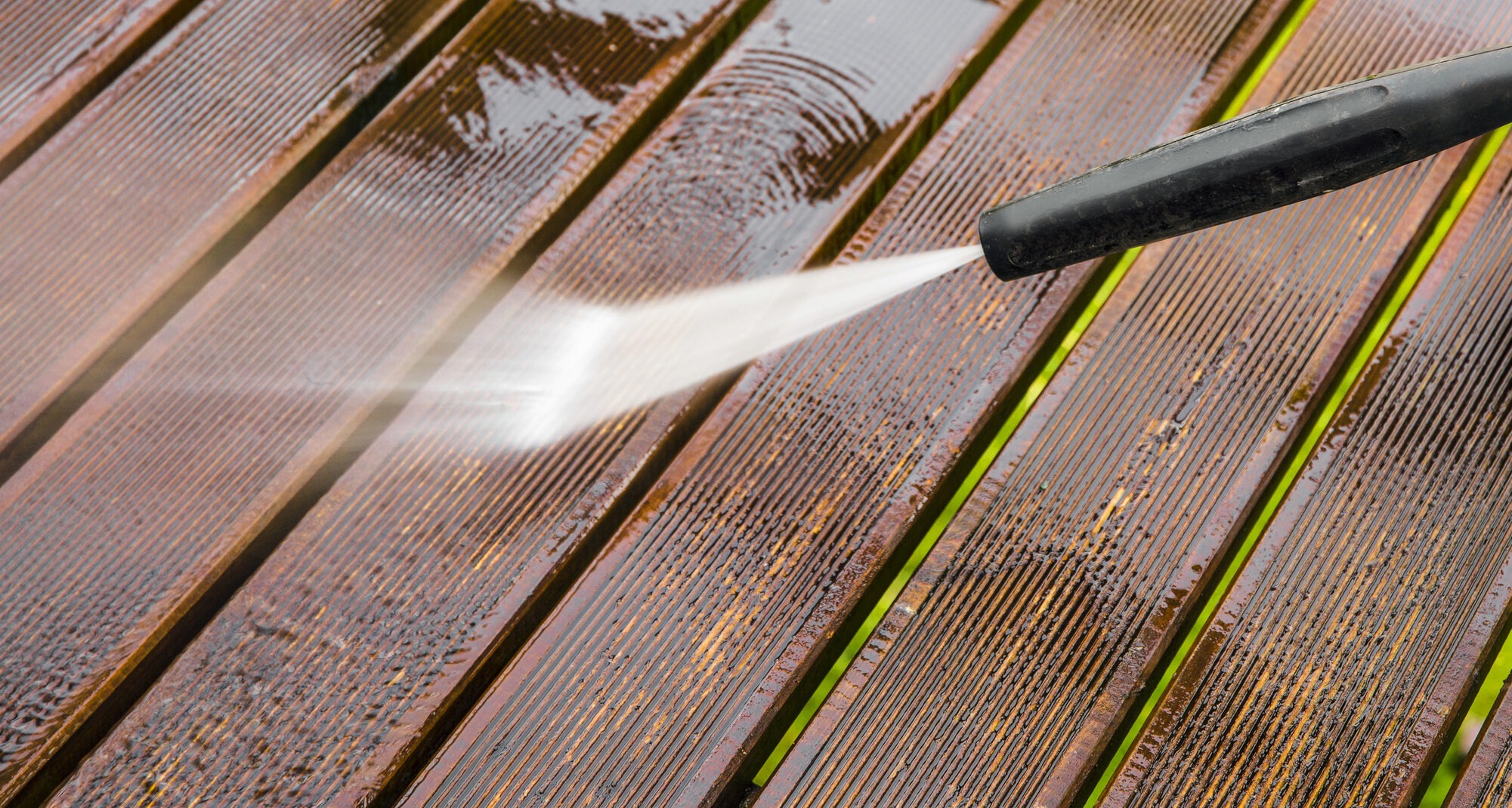 A close-up of a pressure washer being used to clean a wooden deck. The powerful stream of water is blasting away dirt and grime, creating a noticeable difference between the cleaned and uncleaned sections. The deck has narrow gaps between the wooden planks.