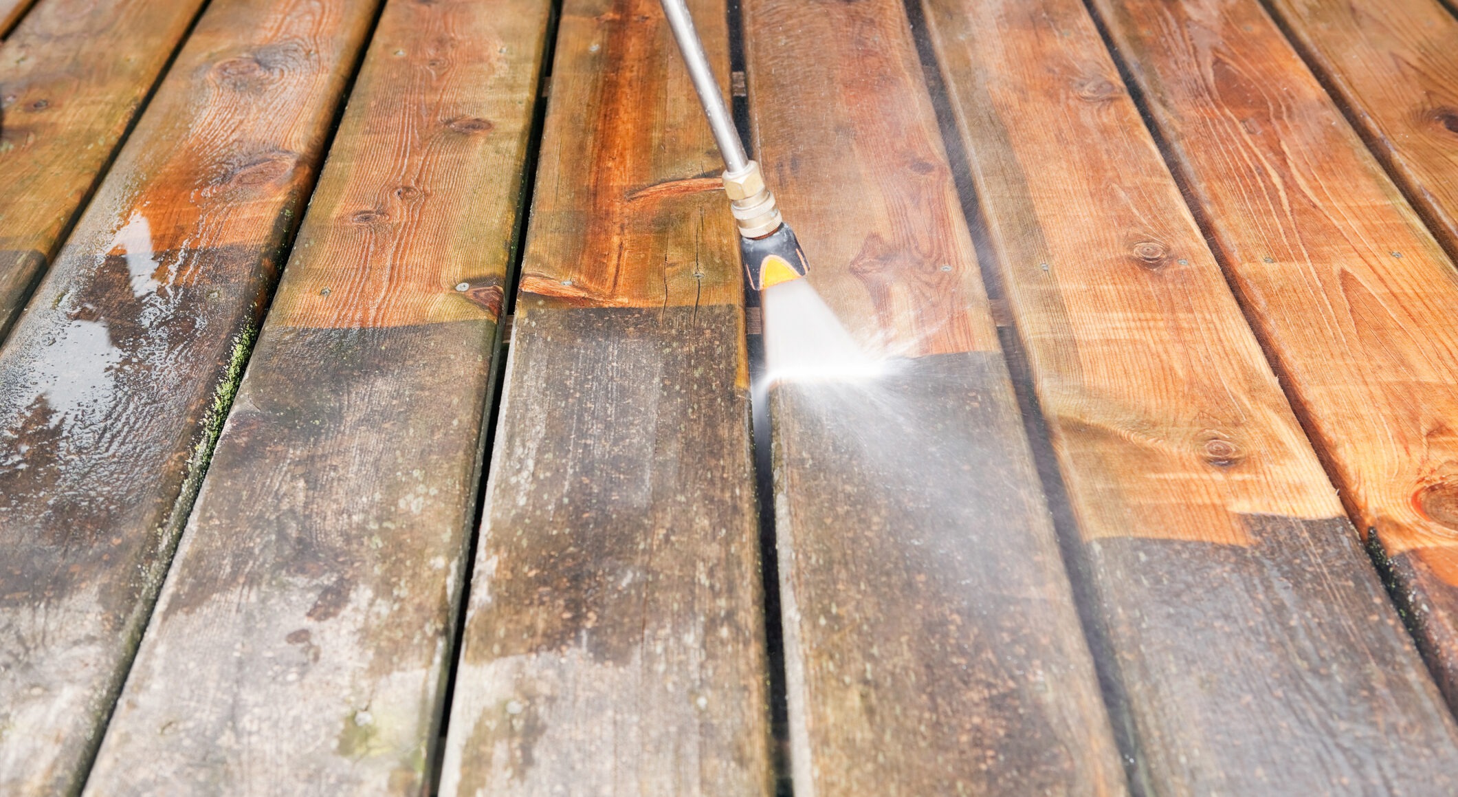 A close-up view of a pressure washer being used to clean a wooden deck. The nozzle emits a powerful jet of water, visibly brightening and removing dirt from the surface, creating a clear contrast between the cleaned and uncleaned sections.