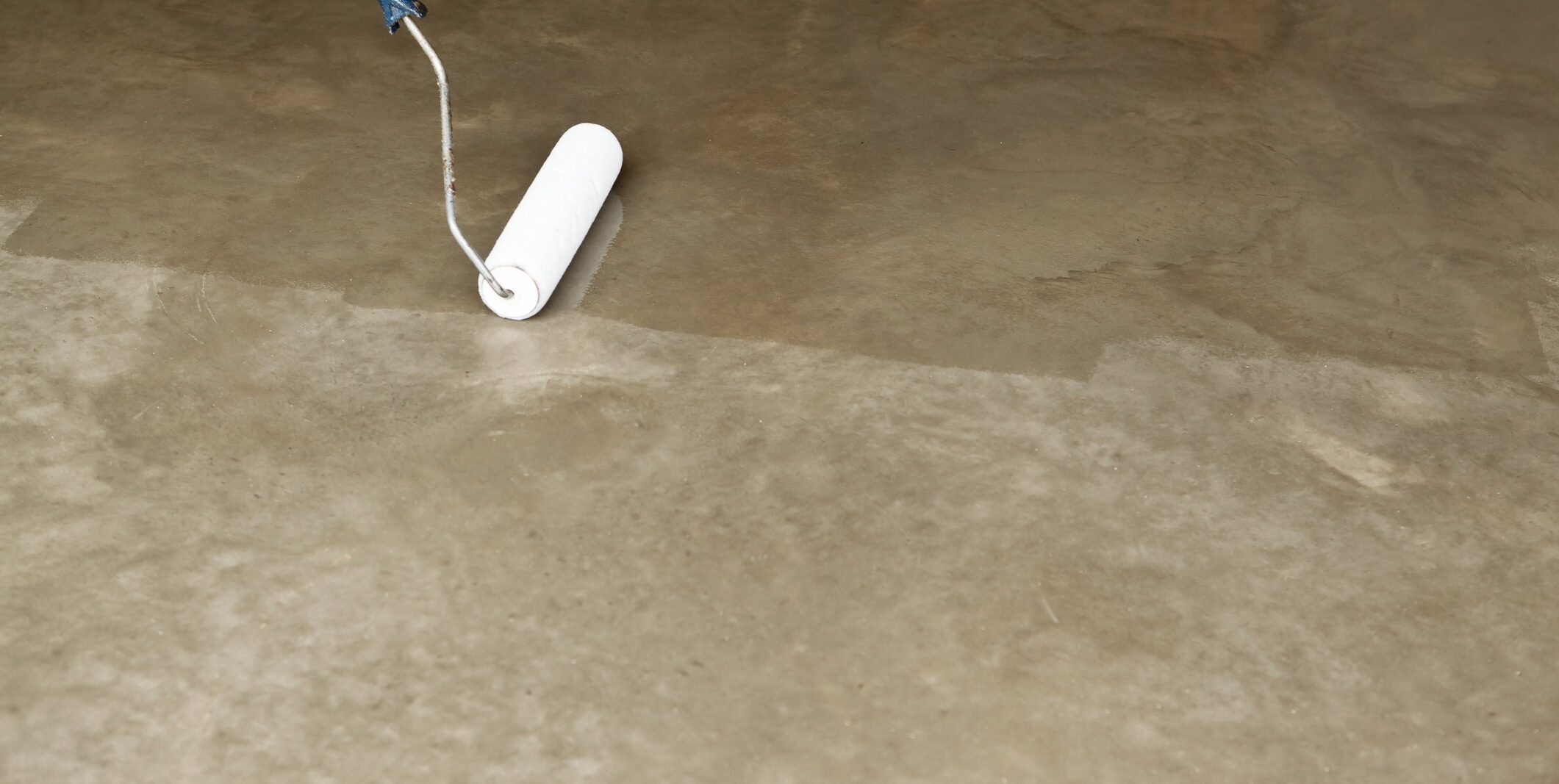 A close-up image shows a person using a white paint roller to apply primer or paint on a smooth concrete floor. The section being painted appears wet and has a darker shade compared to the already dried and lighter area. Only the roller and part of the handle are visible.