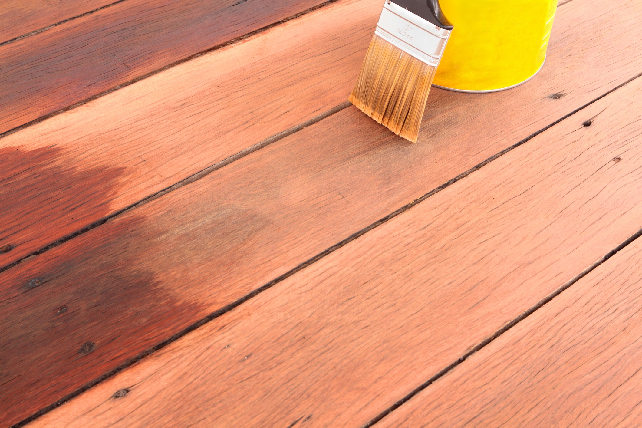 A partially stained wooden deck with a paintbrush and a yellow can of wood stain resting on it. The left side of the planks is darker, indicating the freshly applied stain, while the right side remains unstained and lighter in color.