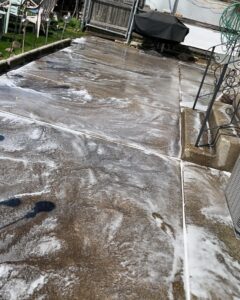 A concrete patio is covered in soapy water, likely being cleaned. Patio furniture and a grill are visible in the background. There are ornamental metal stands on one side, and some steps lead up to the patio. The sun is shining, creating shadows on the wet surface.