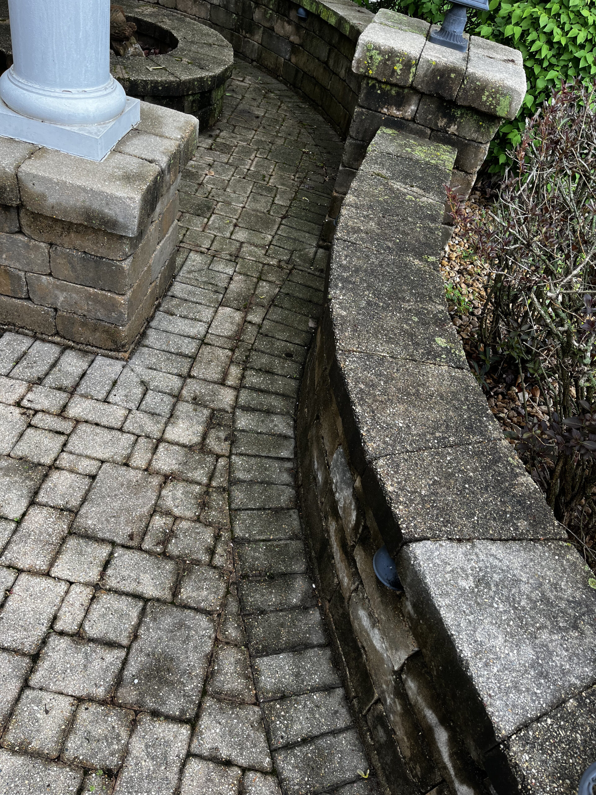 A curved stone path bordered by a low stone wall leads to a circular patio area. The stone work is weathered and damp, with moss growing on the wall. The patio includes a central column. Bushes and greenery are visible on the right side.