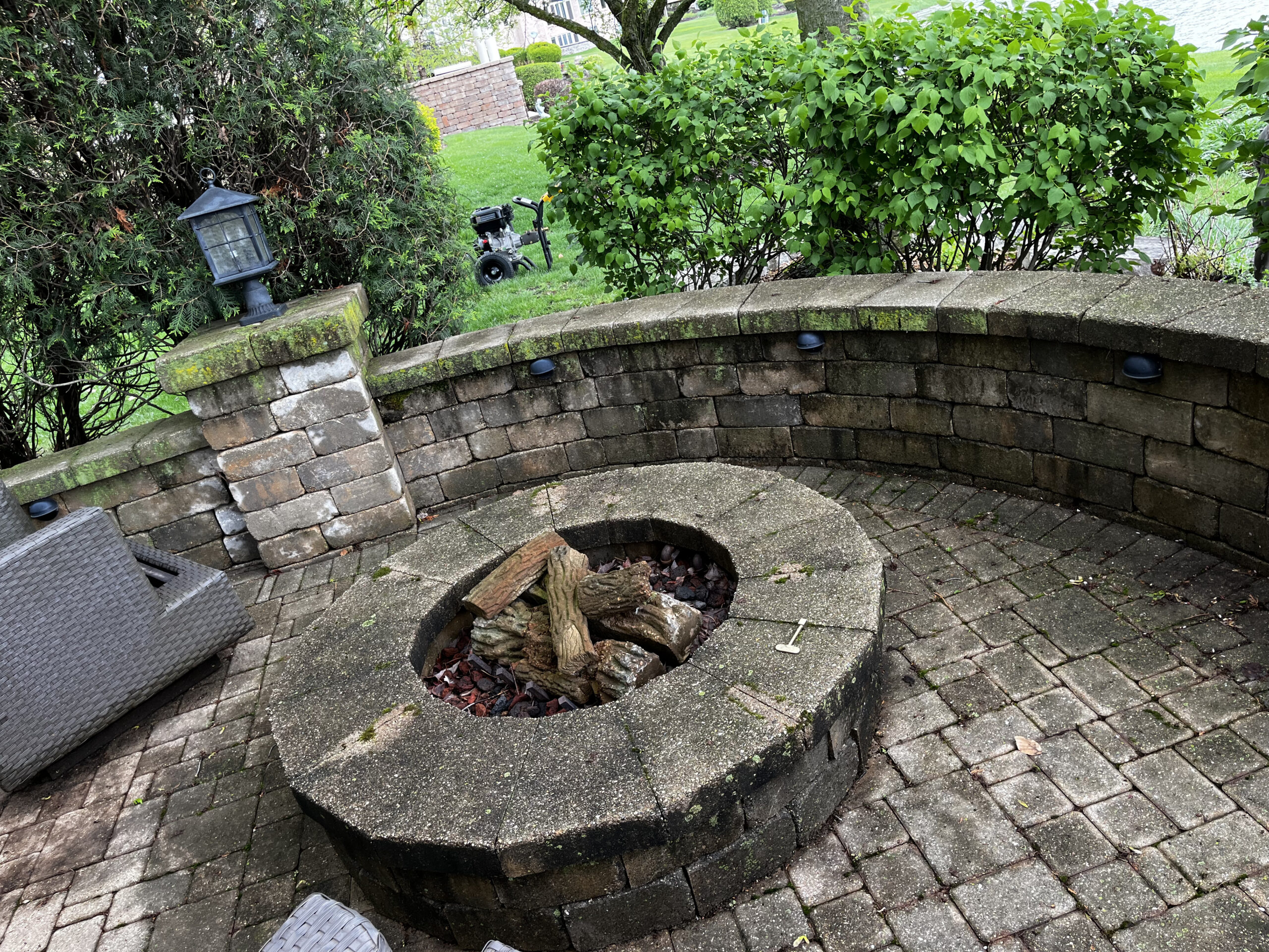 A circular stone fire pit with a wooden log in the center is surrounded by a curved brick seating wall. The patio is paved with brick, and lush green bushes and trees are visible in the background. There are outdoor chairs partially visible on the left.