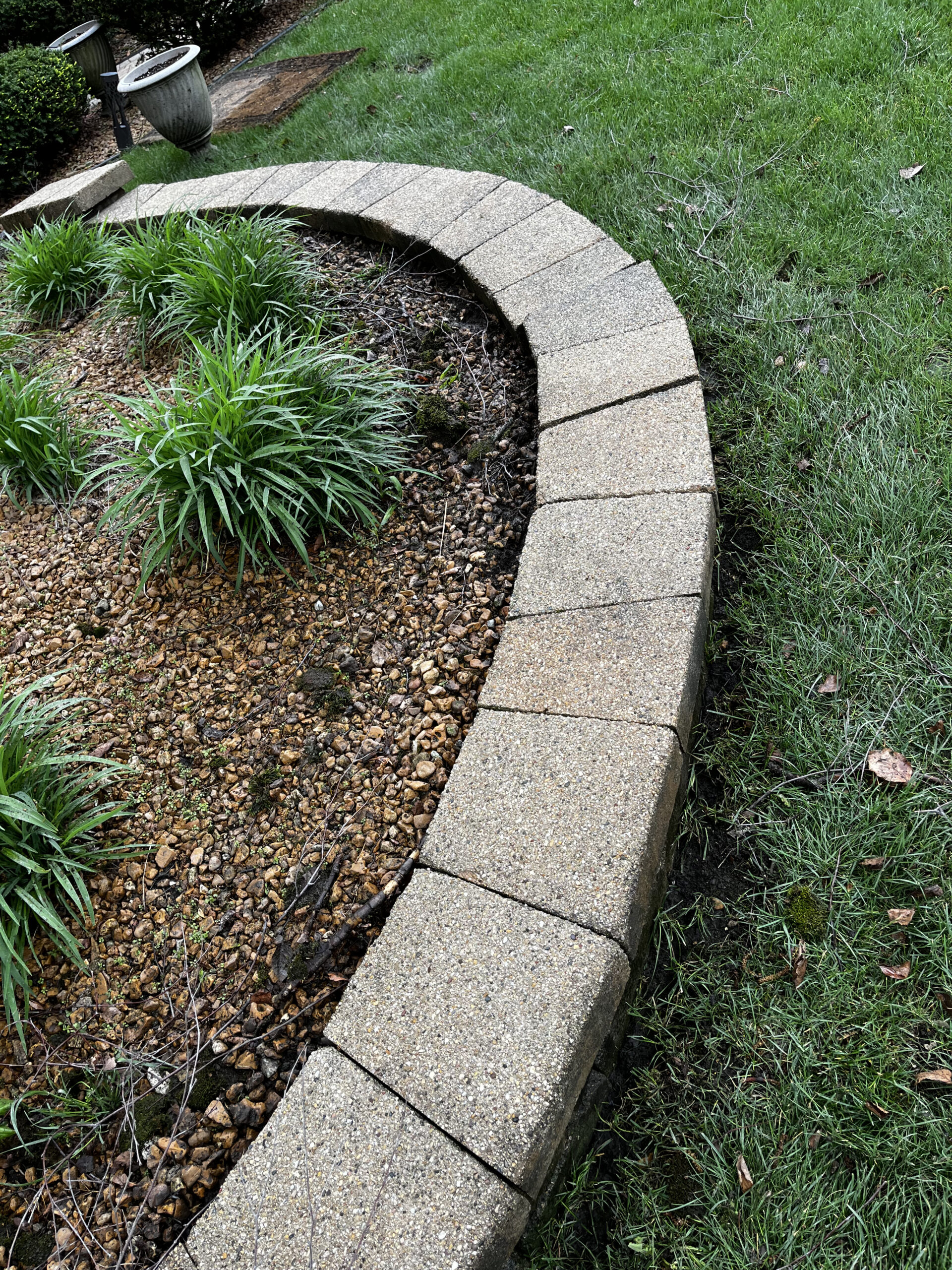 A curved stone border separates a small garden bed filled with green plants and gravel from the surrounding grassy lawn. A decorative pot sits at the end of the border in the background.