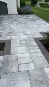 A neatly arranged patio with large gray pavers forming an intricate, modern pattern. The design includes dark gray border pavers. Adjacent areas have garden beds with soil and trimmed green plants. A grassy lawn and part of a white brick building are visible in the background.