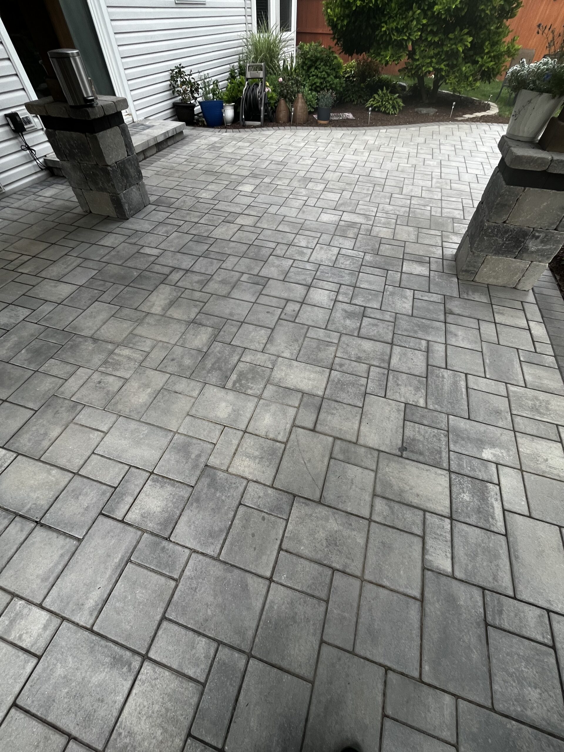 A newly laid, clean, stone patio with interlocking gray rectangular pavers. The patio is adjacent to a white siding house with two stone columns supporting a small roof. Potted plants and a garden hose are visible in the background near a green bush.